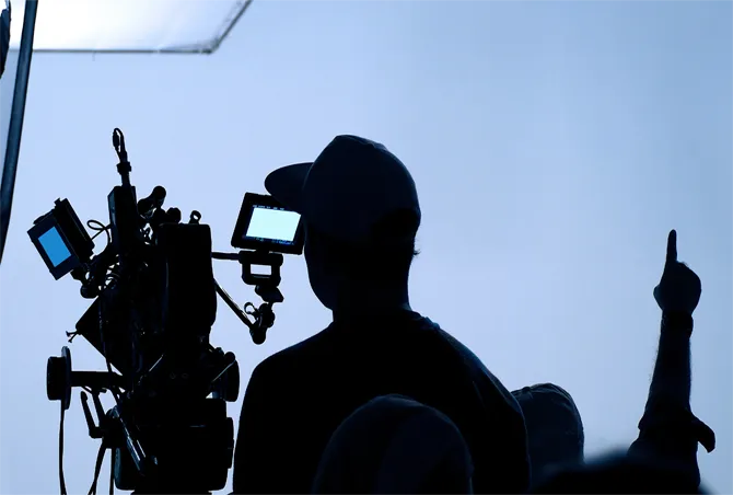 Silhouette of a person operating a film camera with monitors, backlit by soft lighting, indicating a film production environment. tv production studio mainstream ent. 